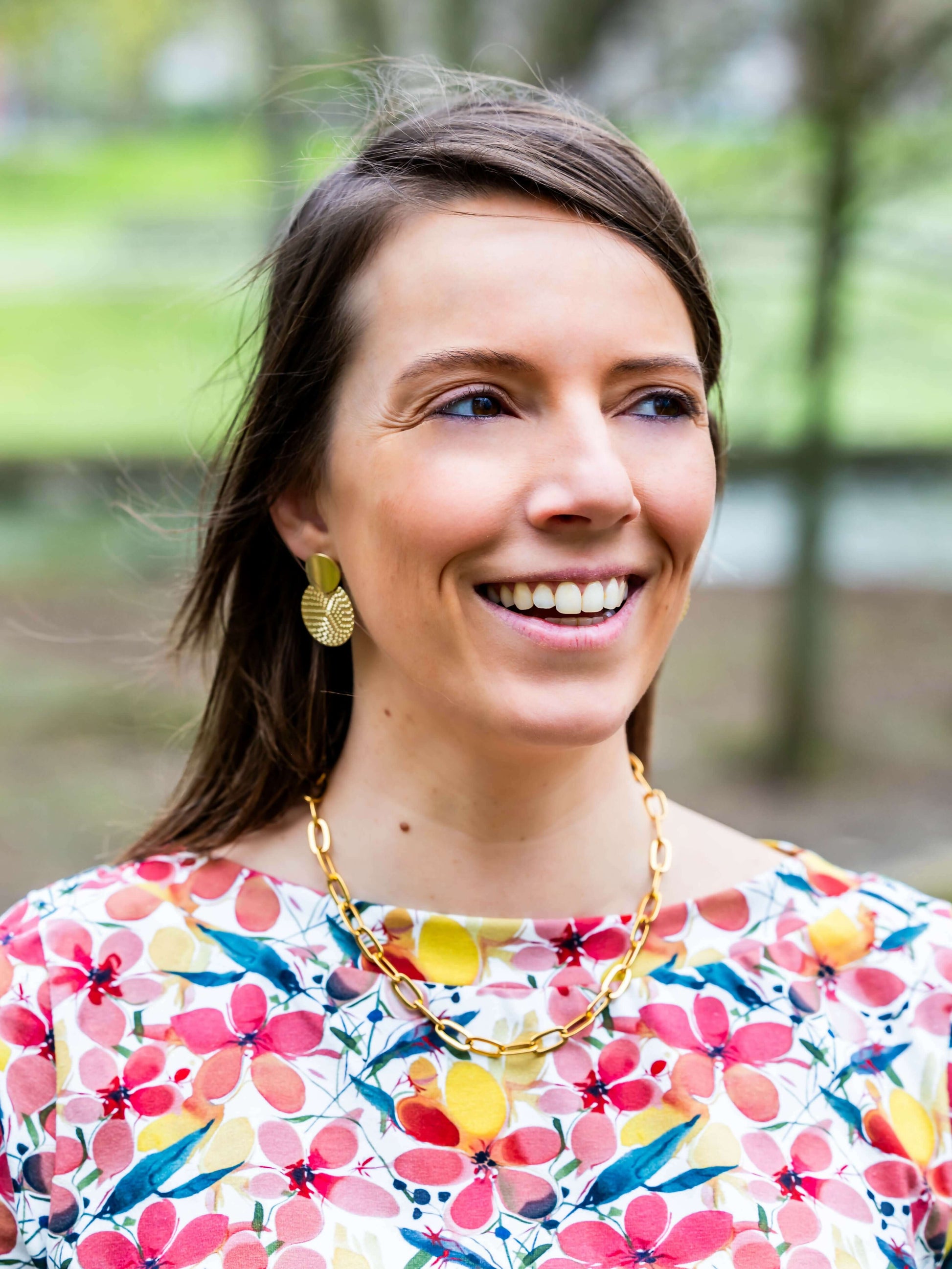 Femme souriante portant un collier doré et un chemisier à motifs colorés, dans un parc.