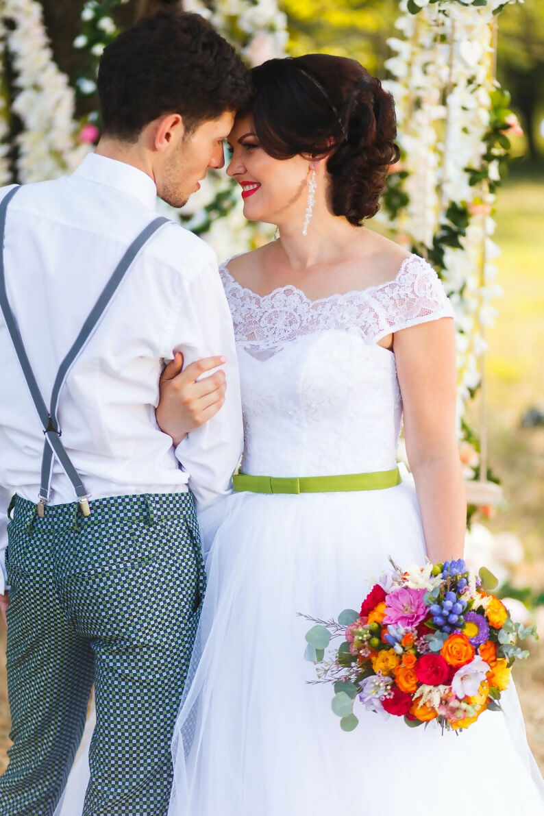Couple de mariés élégants sous une arche florale, robe blanche et bouquet coloré en été