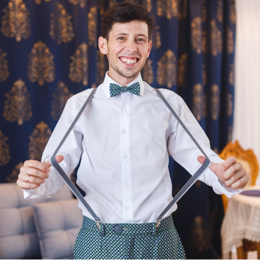 Homme souriant portant une chemise blanche avec des bretelles et un nœud papillon, devant un fond élégant de rideaux décoratifs.