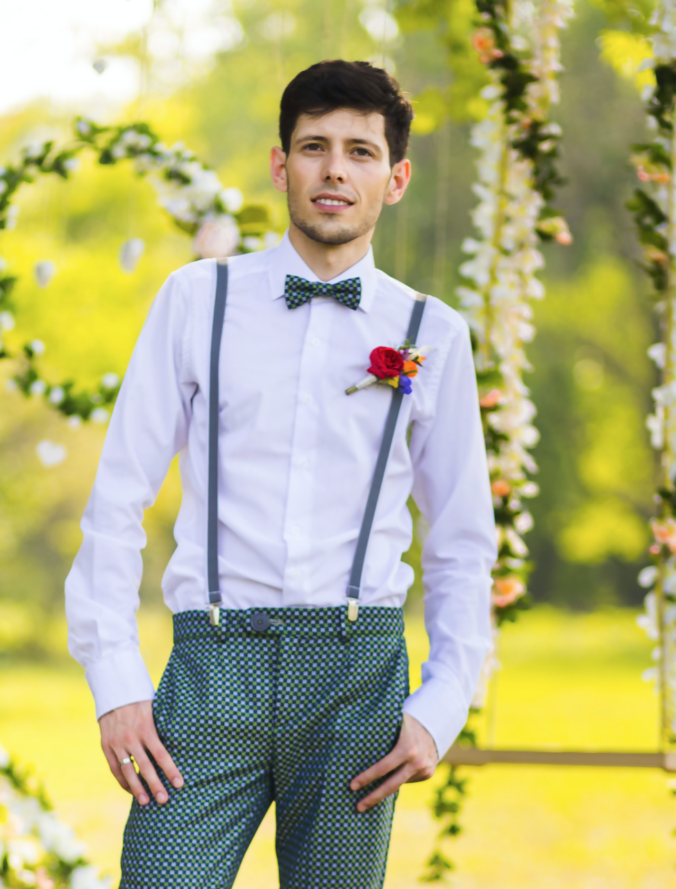 Homme élégant vêtu d'une chemise blanche avec bretelles, debout devant une arche florale, style mariage en extérieur.