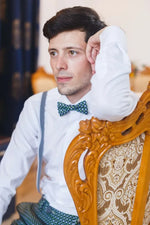 Homme assis sur une chaise en bois, portant une chemise blanche et un nœud papillon bleu, regard pensif.