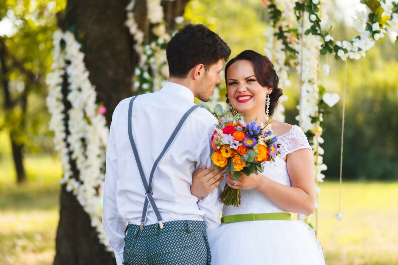 Couple marié heureux posant avec bouquet coloré lors d'une cérémonie de mariage en plein air.