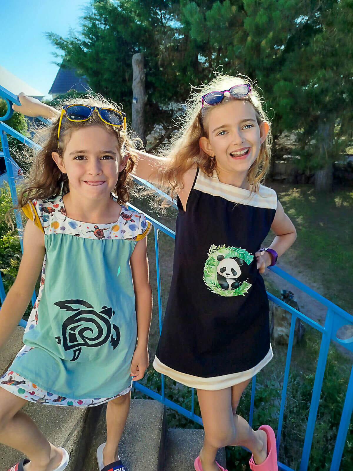 Deux filles portant des robes colorées avec des motifs d'animaux, souriant sur un escalier en extérieur.