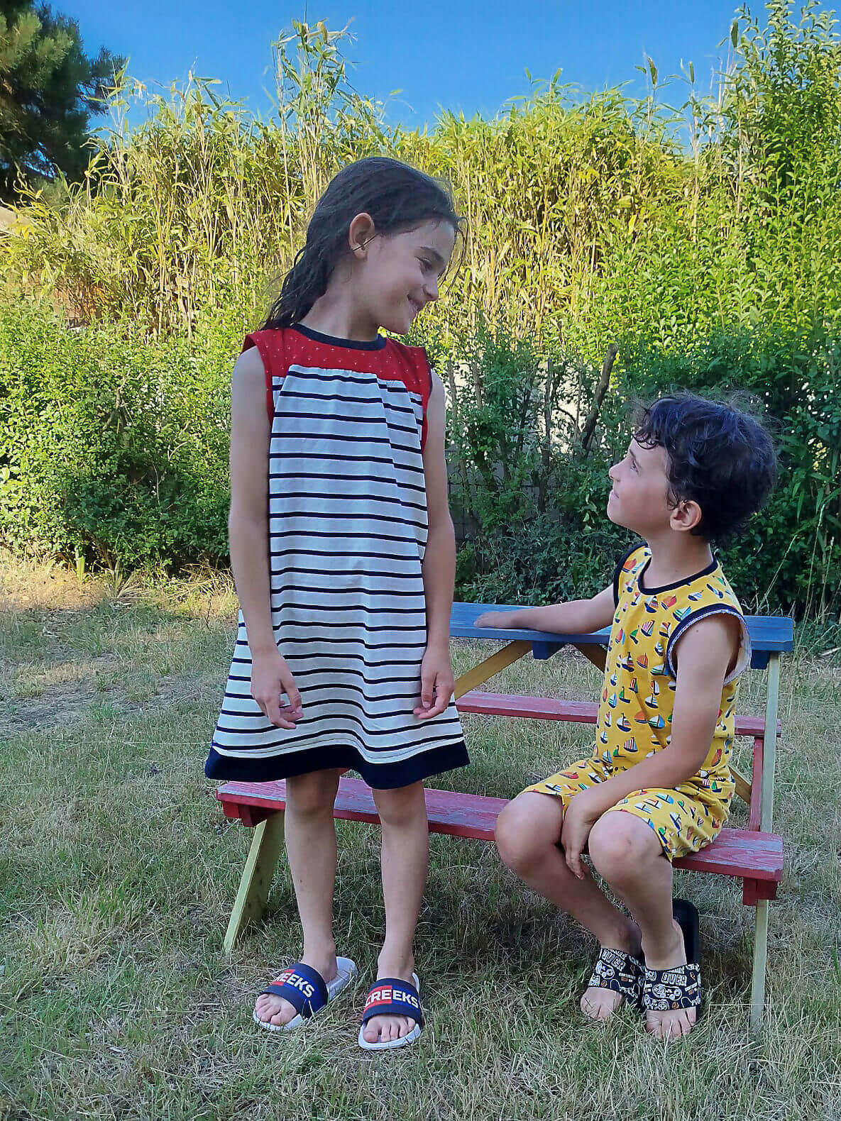 Deux enfants en plein air, une fille en robe rayée rouge et noire et un garçon en tenue jaune, assis sur un banc multicolore.