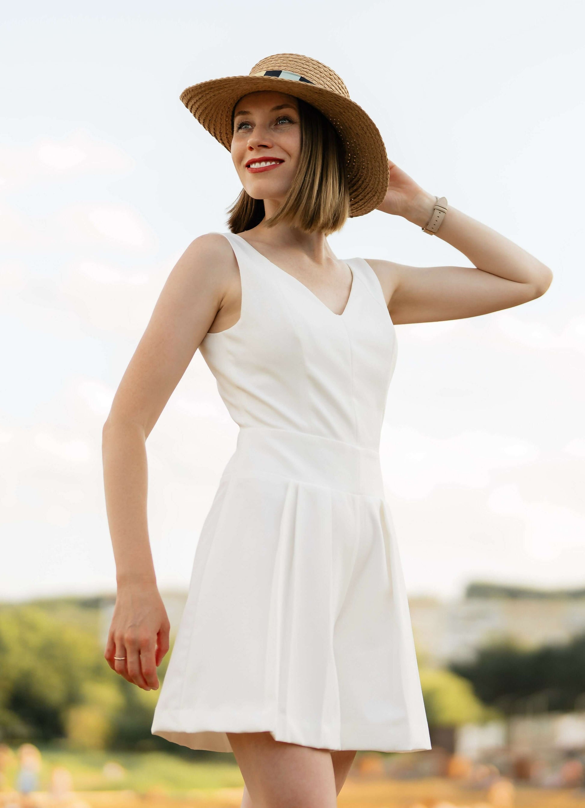 Femme en robe d'été blanche avec chapeau de paille dans un champ, souriante sous un ciel ensoleillé.