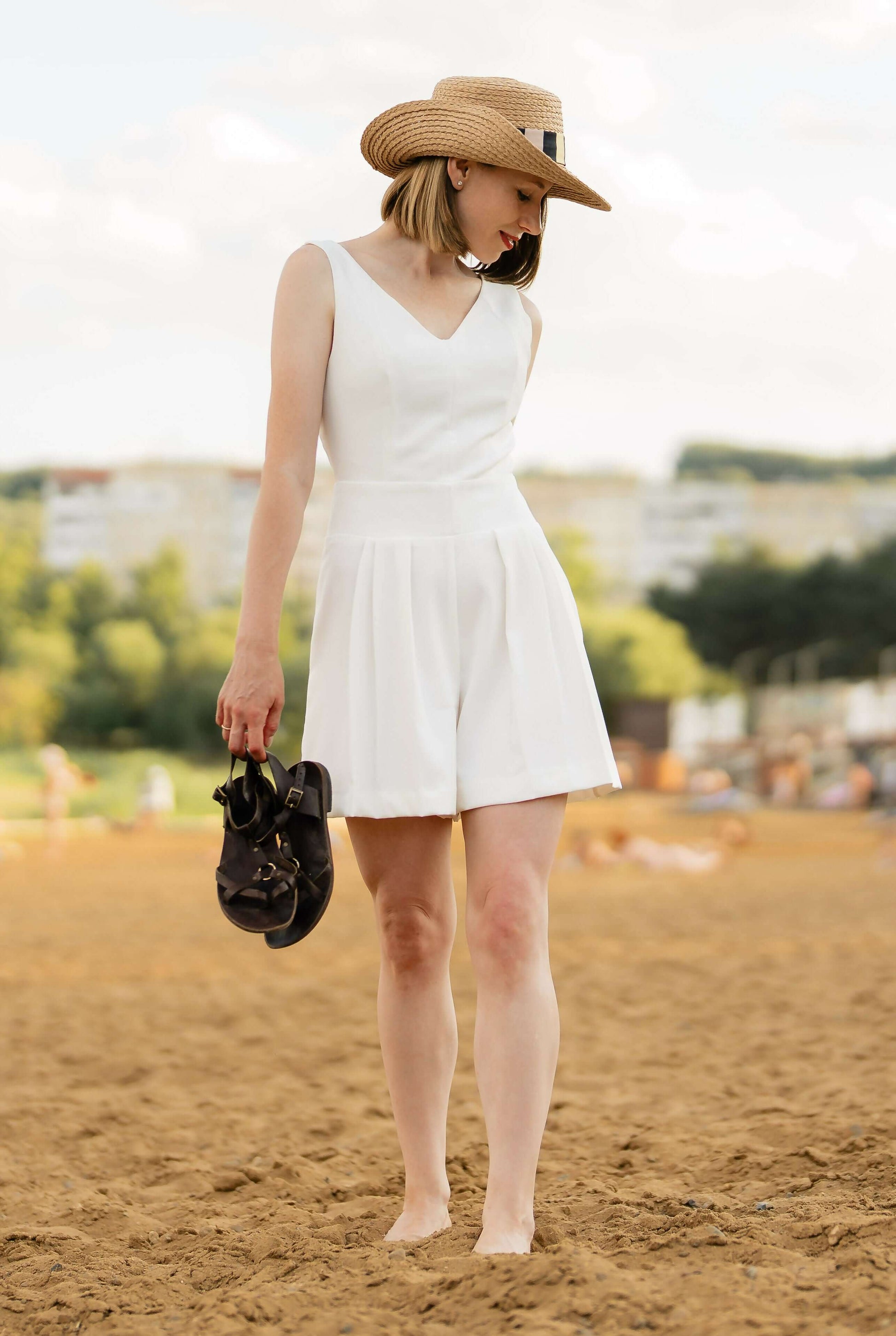 Femme en combinaison blanche élégante tenant des sandales sur une plage, portant un chapeau de paille.