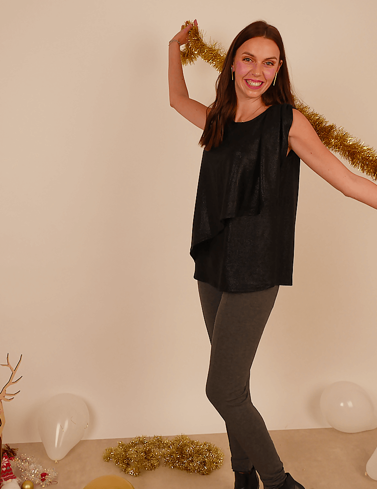 Femme souriante portant le débardeur Cara noir, élégant et décontracté, avec des leggings gris, en arrière-plan festif.