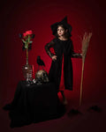 Petite fille en déguisement de sorcière noire avec chapeau, devant une table décorée pour Halloween.