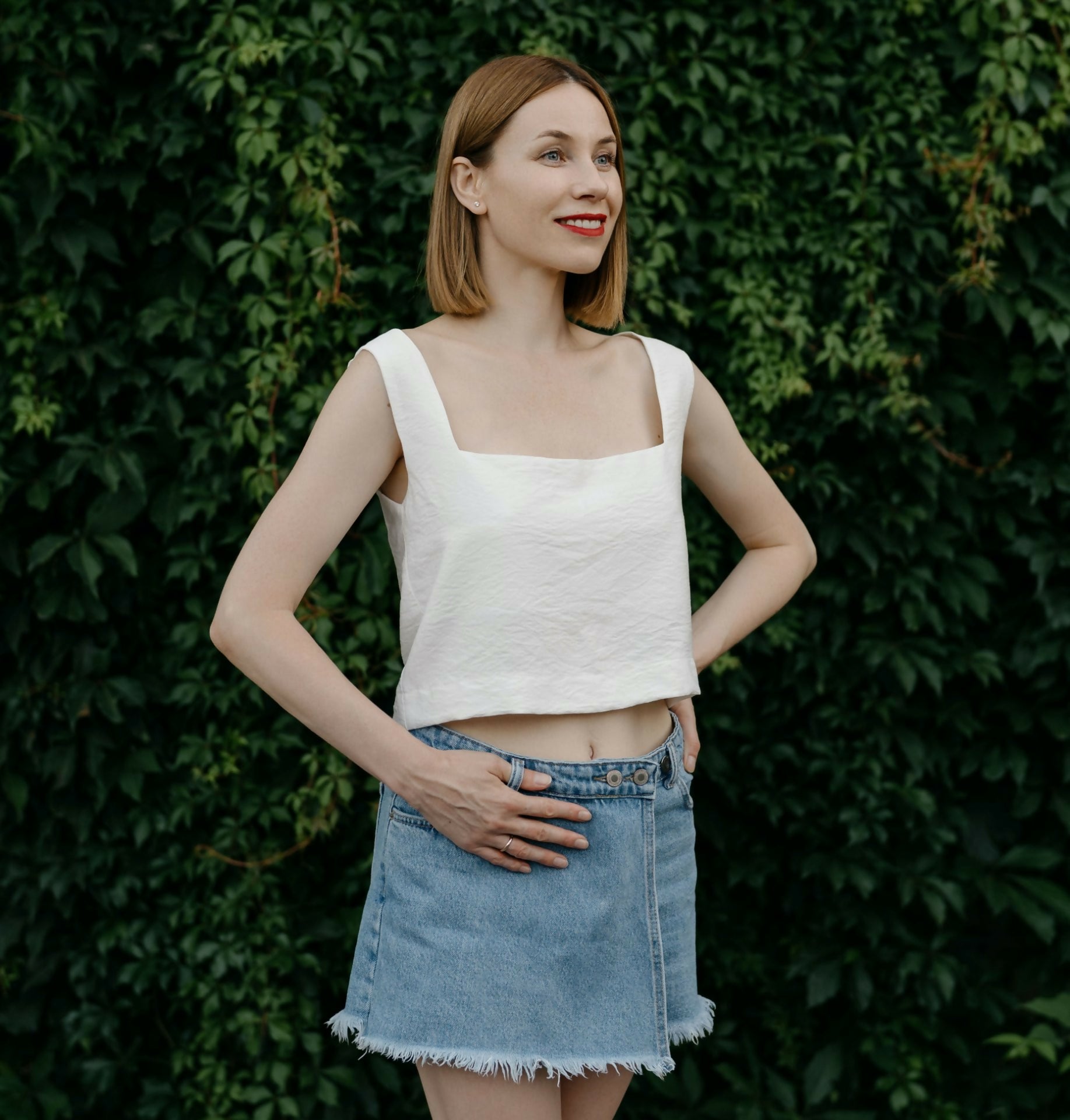 Femme posant en top blanc et jupe en jean devant un fond de végétation luxuriante.