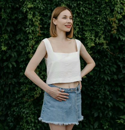 Femme posant en top blanc et jupe en jean devant un fond de végétation luxuriante.