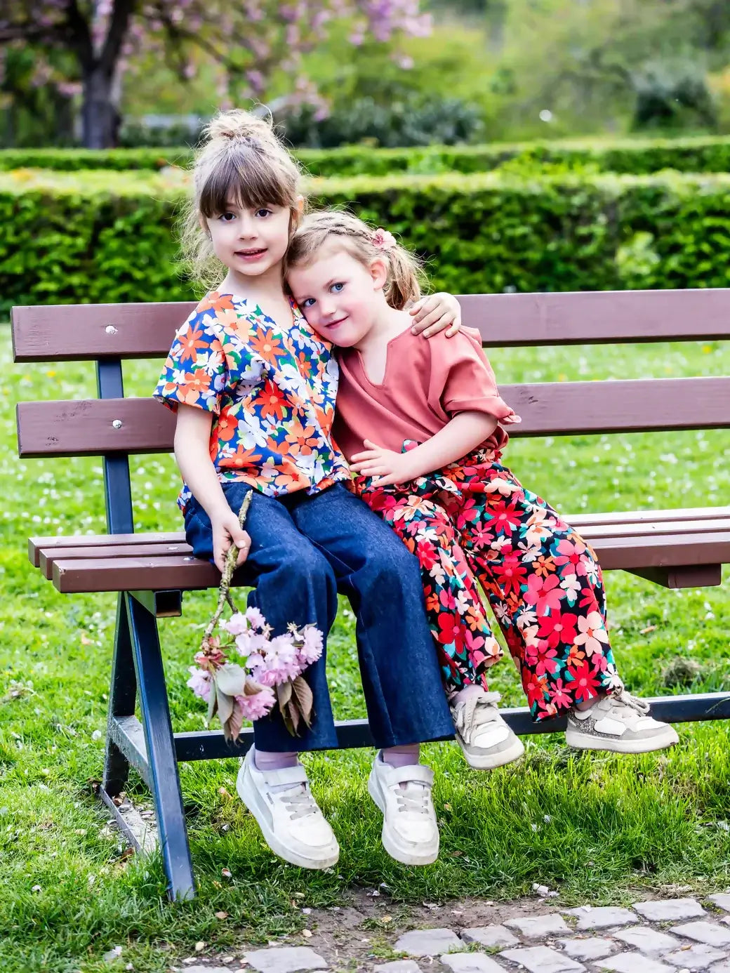Deux enfants assises sur un banc, portant des vêtements colorés avec des motifs floraux, entourées d'un jardin verdoyant en arrière-plan.