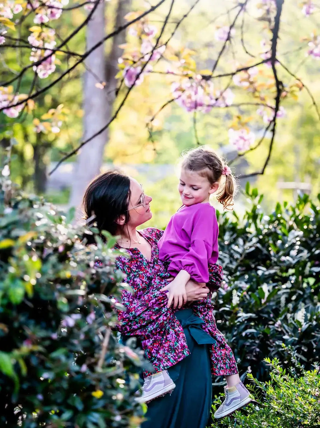Une femme portant une blouse florale tient une petite fille dans un jardin fleuri sous des arbres printaniers.