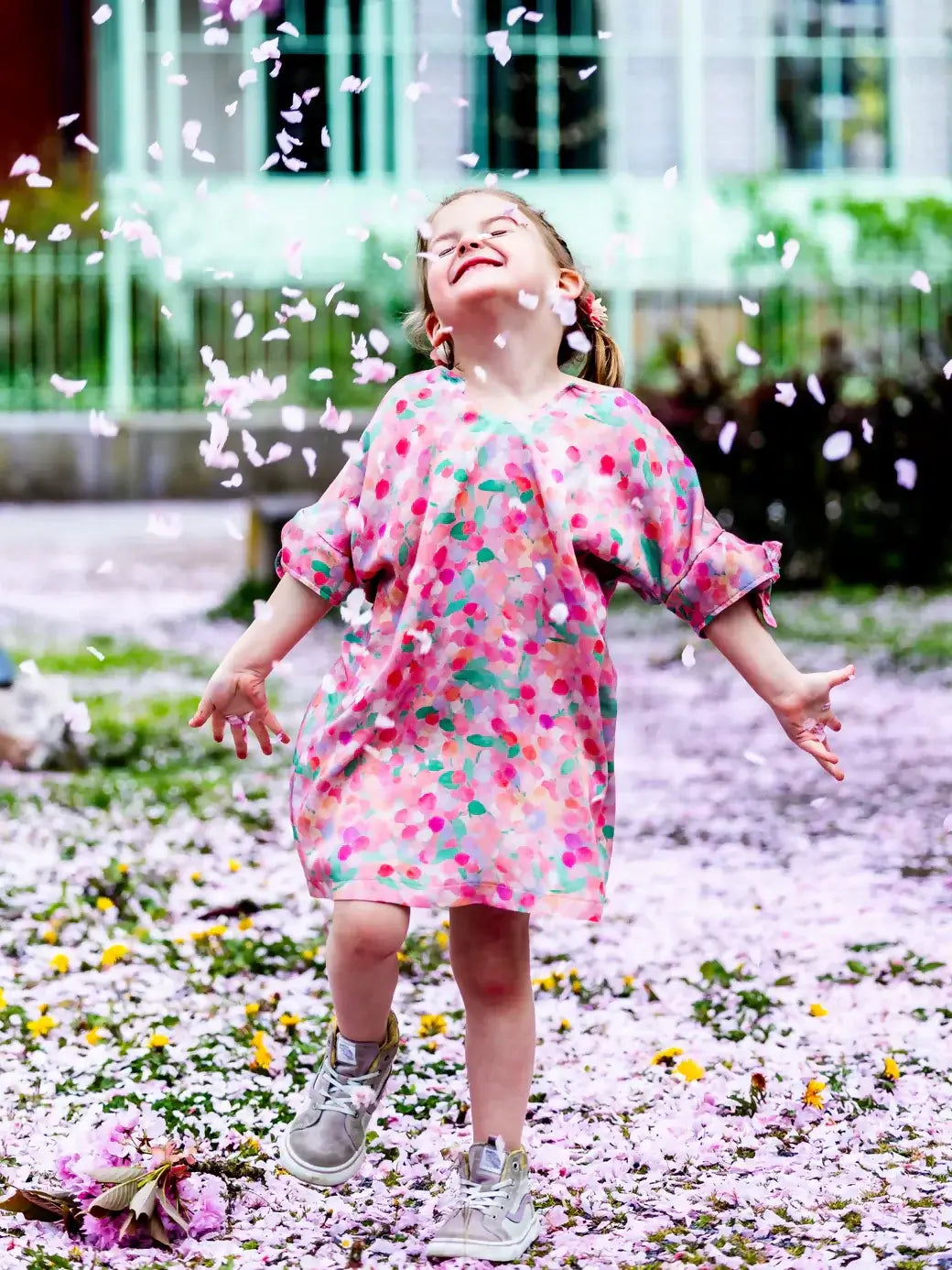 Enfant en robe colorée joyeuse parmi des pétales de fleurs tombants dans un parc, capturant un moment de bonheur insouciant.