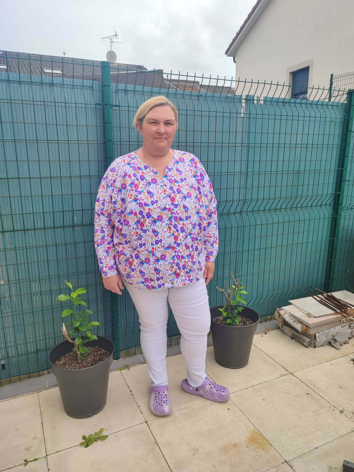 Person wearing a floral blouse and white pants standing on a patio with potted plants.