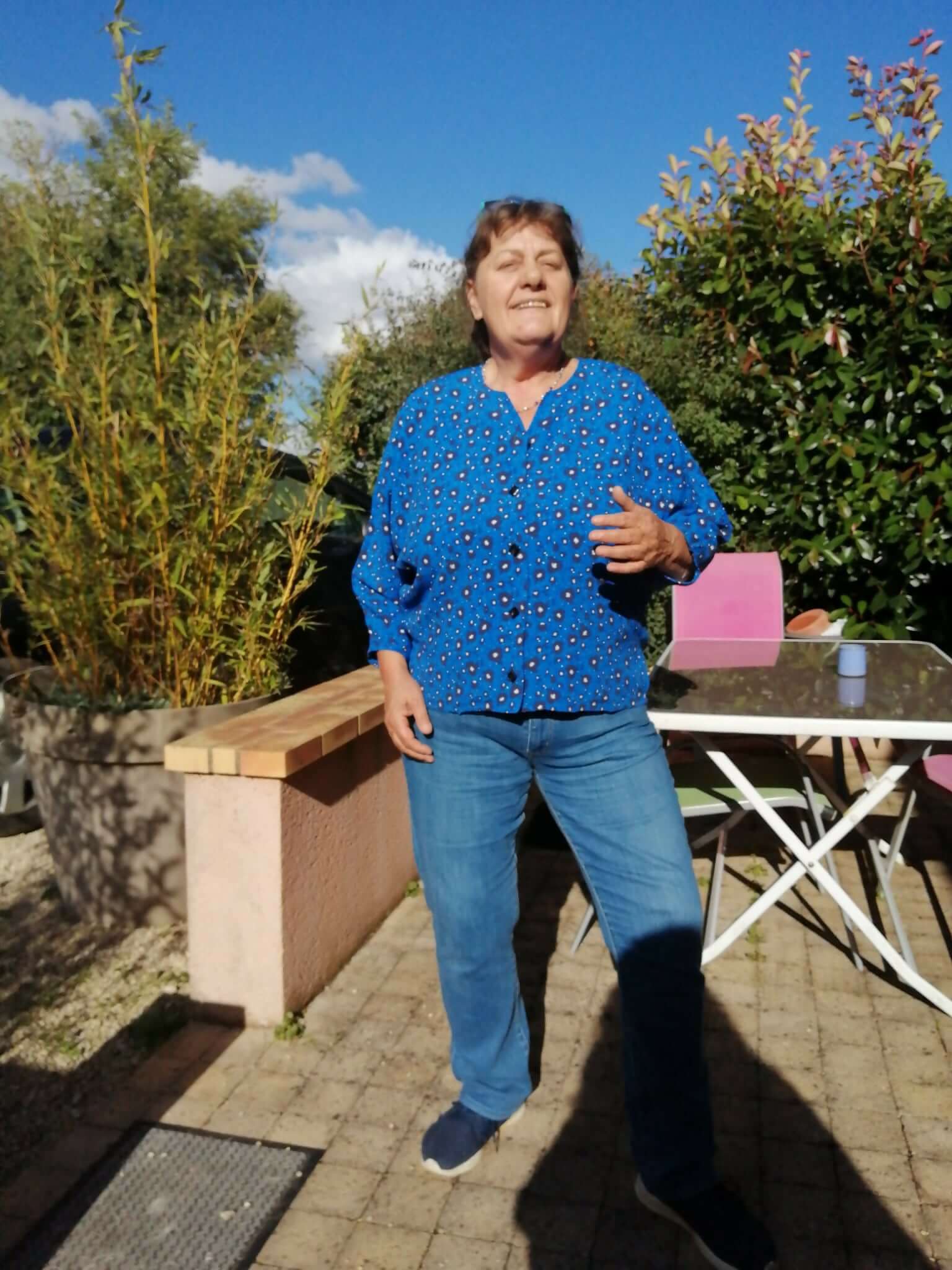 Personne posant en extérieur, portant une chemise bleue avec motifs. Soleil et végétation en arrière-plan.