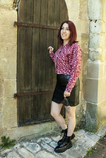 Femme en jupe noire et chemisier floral devant une porte en bois rustique.