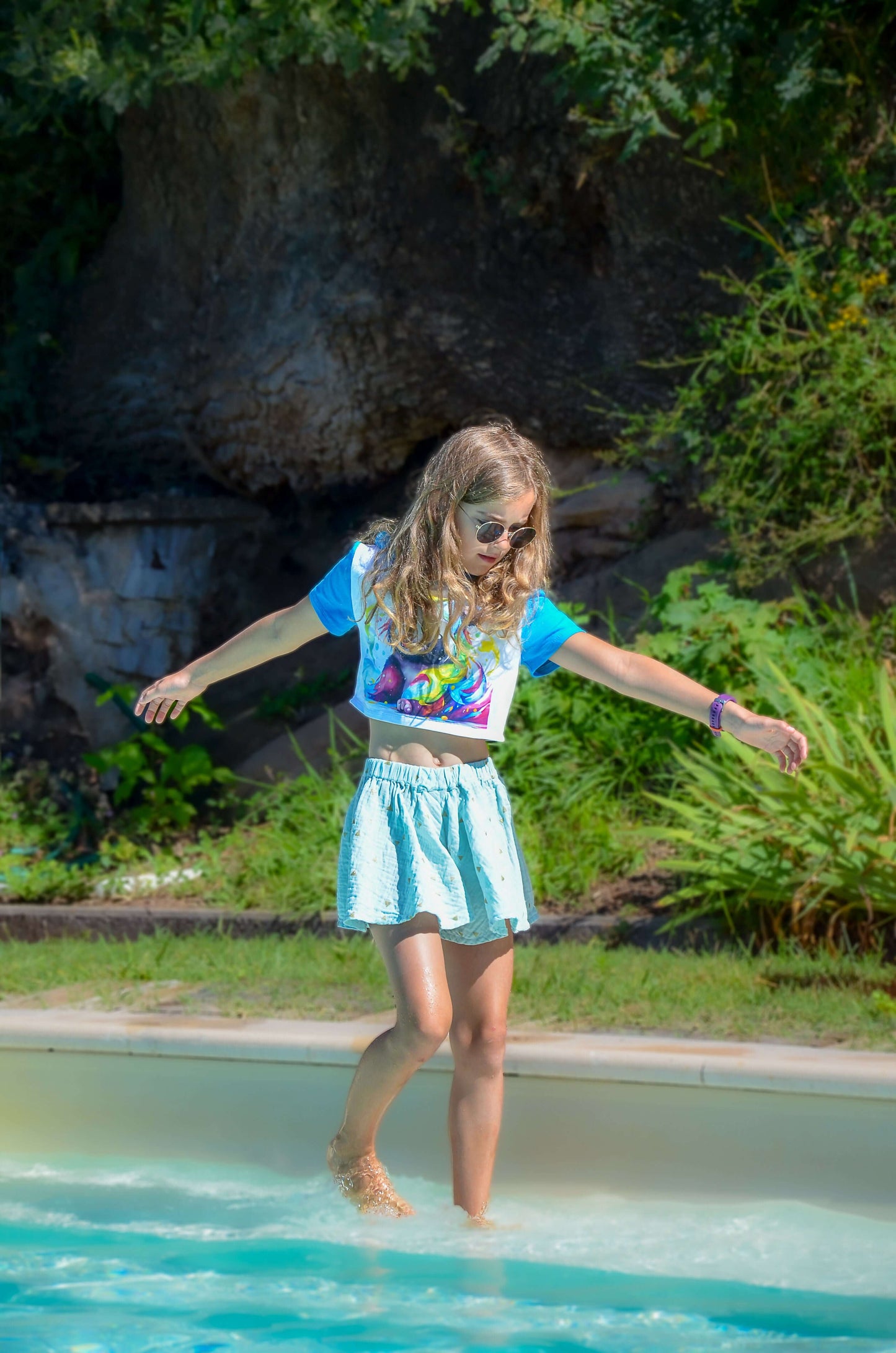 Young girl in a colorful outfit, playfully walking in shallow water at a sunny outdoor setting.
