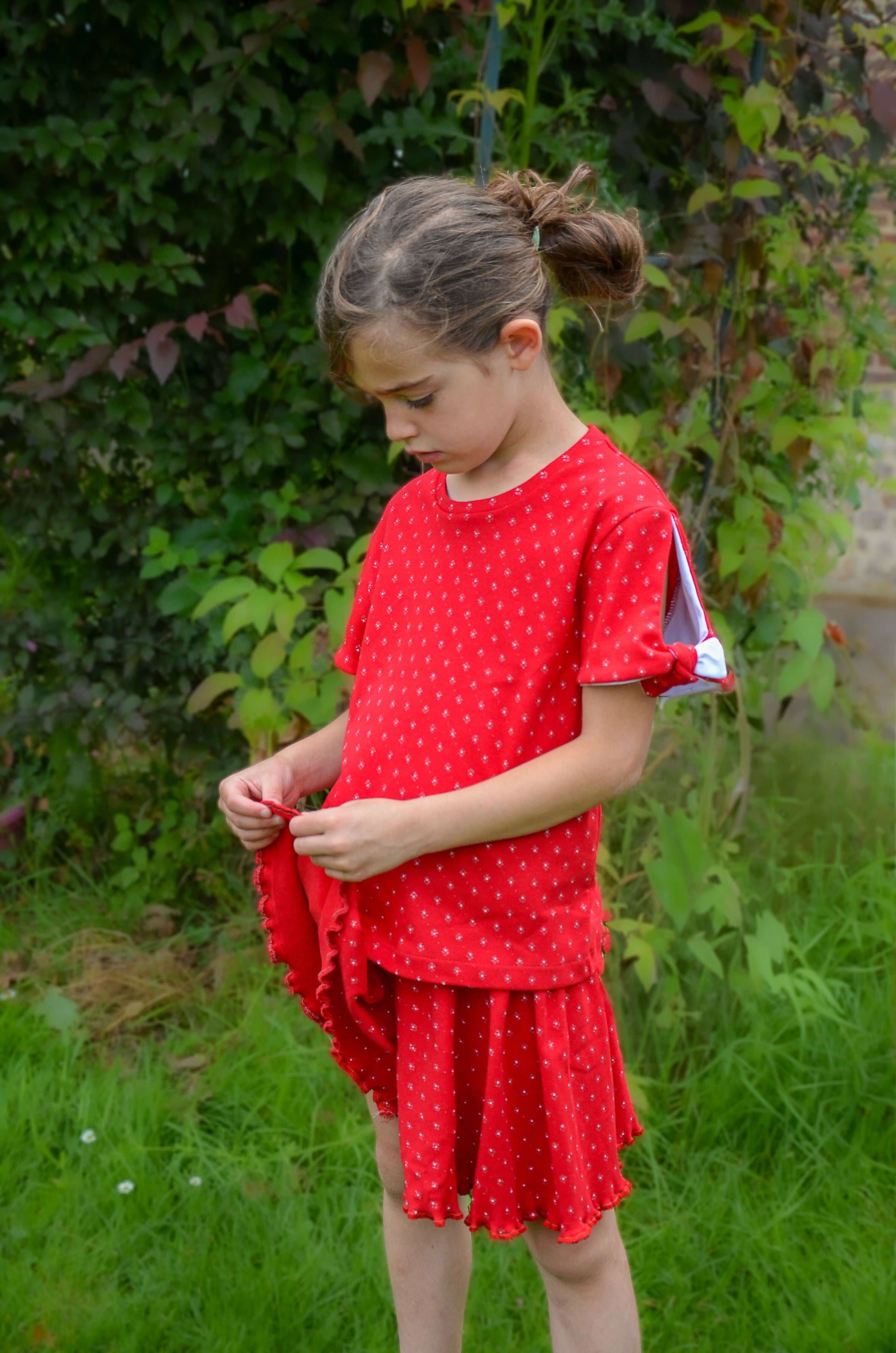 Enfant portant un jupe-short rouge à pois, souriant dans un jardin verdoyant.