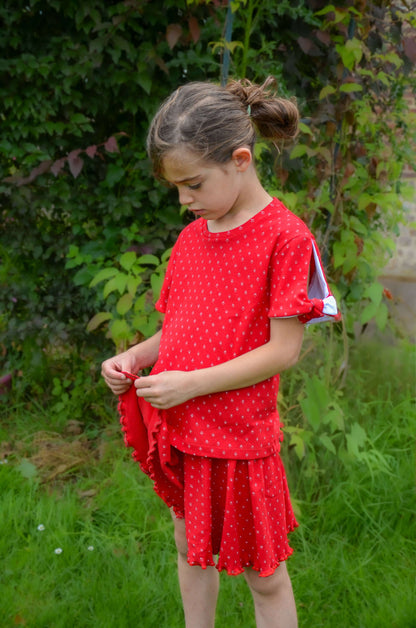 Enfant portant un jupe-short rouge à pois, souriant dans un jardin verdoyant.