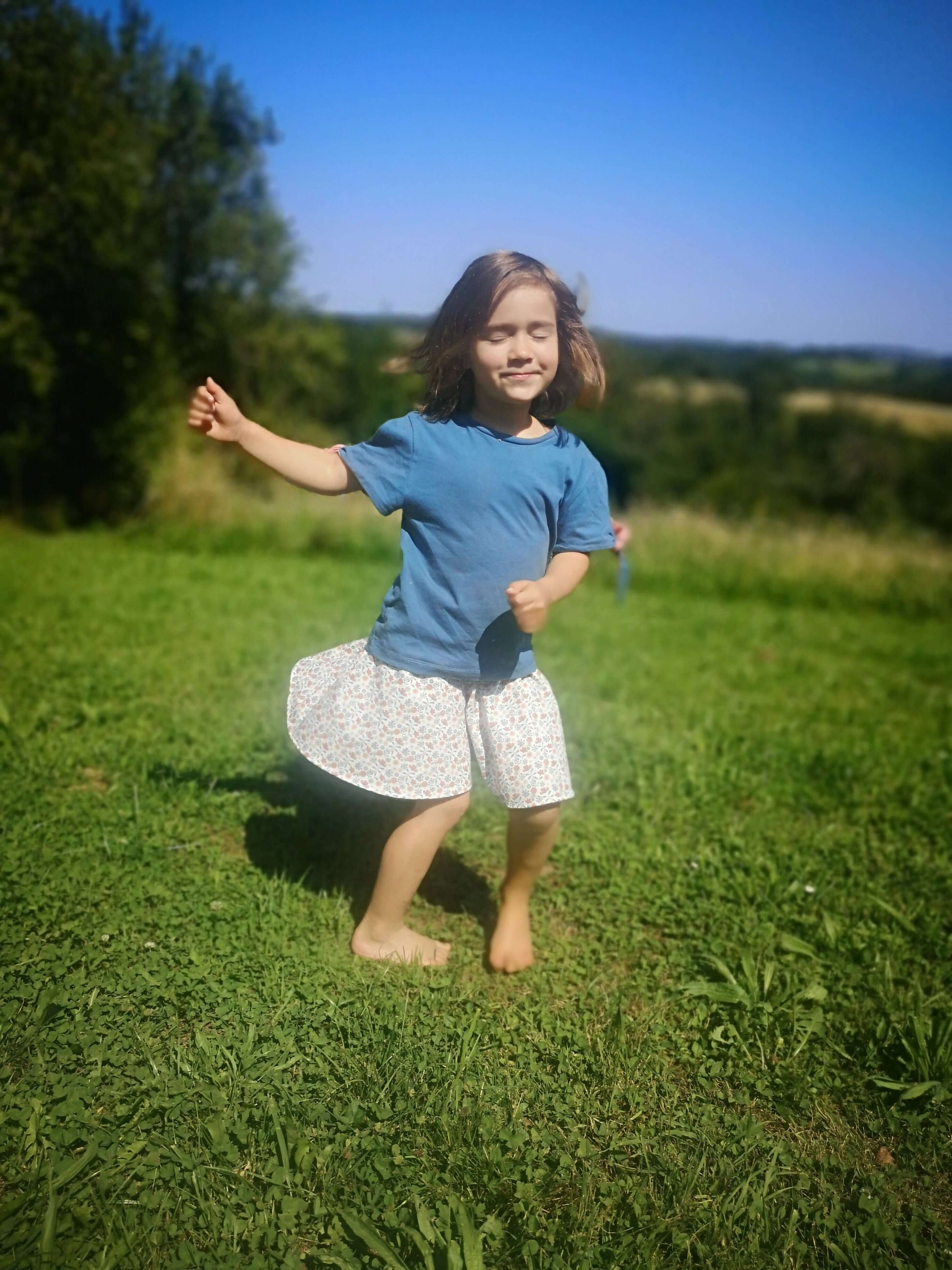 Enfant souriant portant un jupe-short à fleurs et un t-shirt, sautant joyeusement dans un champ ensoleillé.