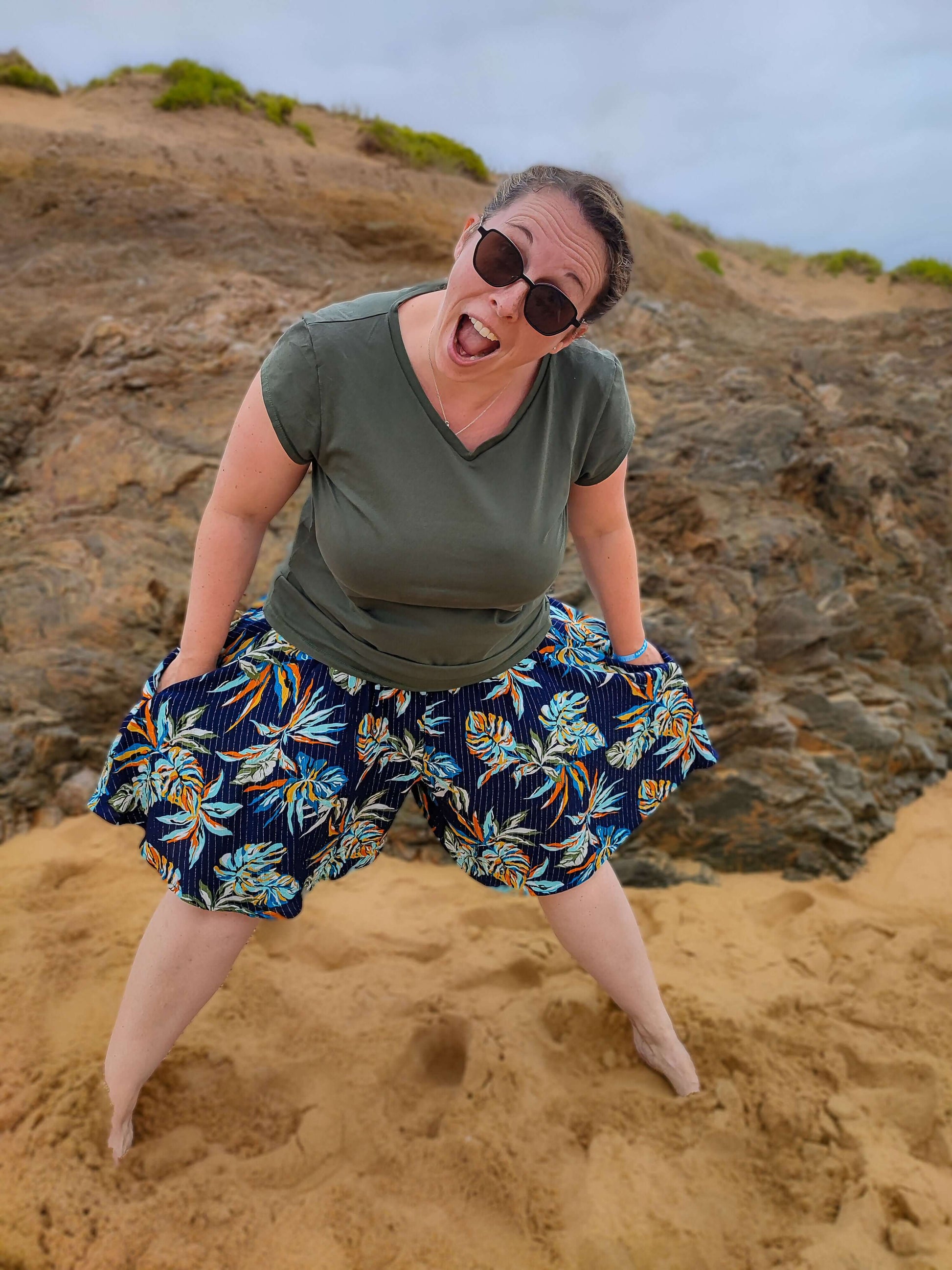 Femme souriante portant un short à fleurs sur la plage, avec des lunettes de soleil et un t-shirt vert.