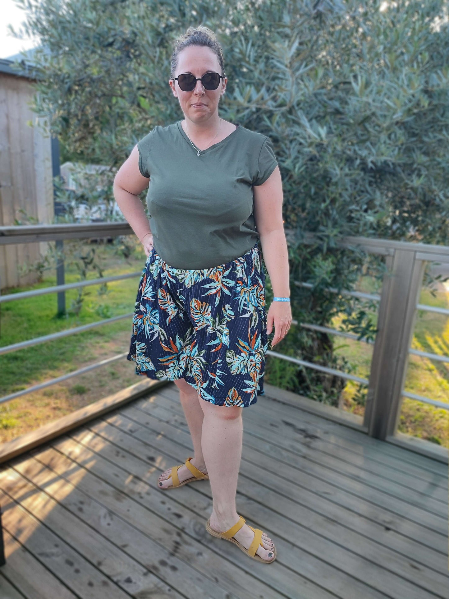 Femme portant un jupe-short floral sur une terrasse, avec un t-shirt vert et lunettes de soleil, idéale pour l'été.