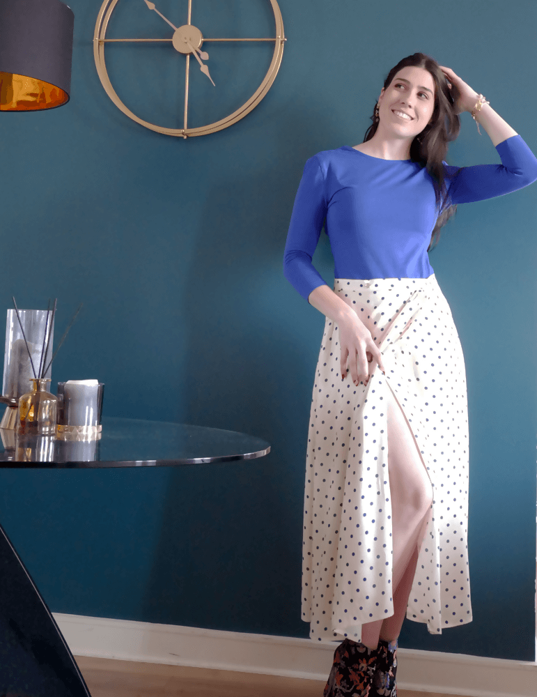 Femme souriante portant la jupe Suzie à pois, style chic et décontracté devant un mur bleu.