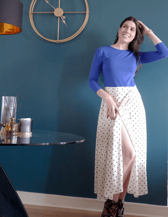 Femme souriante portant la jupe Suzie à pois, style chic et décontracté devant un mur bleu.