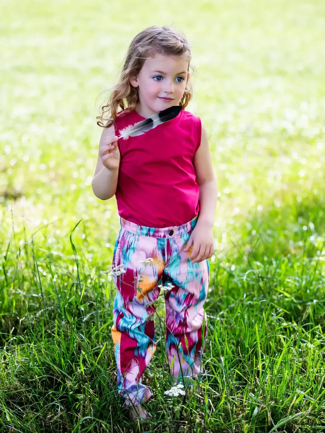 Enfant portant un pantalon coloré dans un parc, tenant une plume, en été.