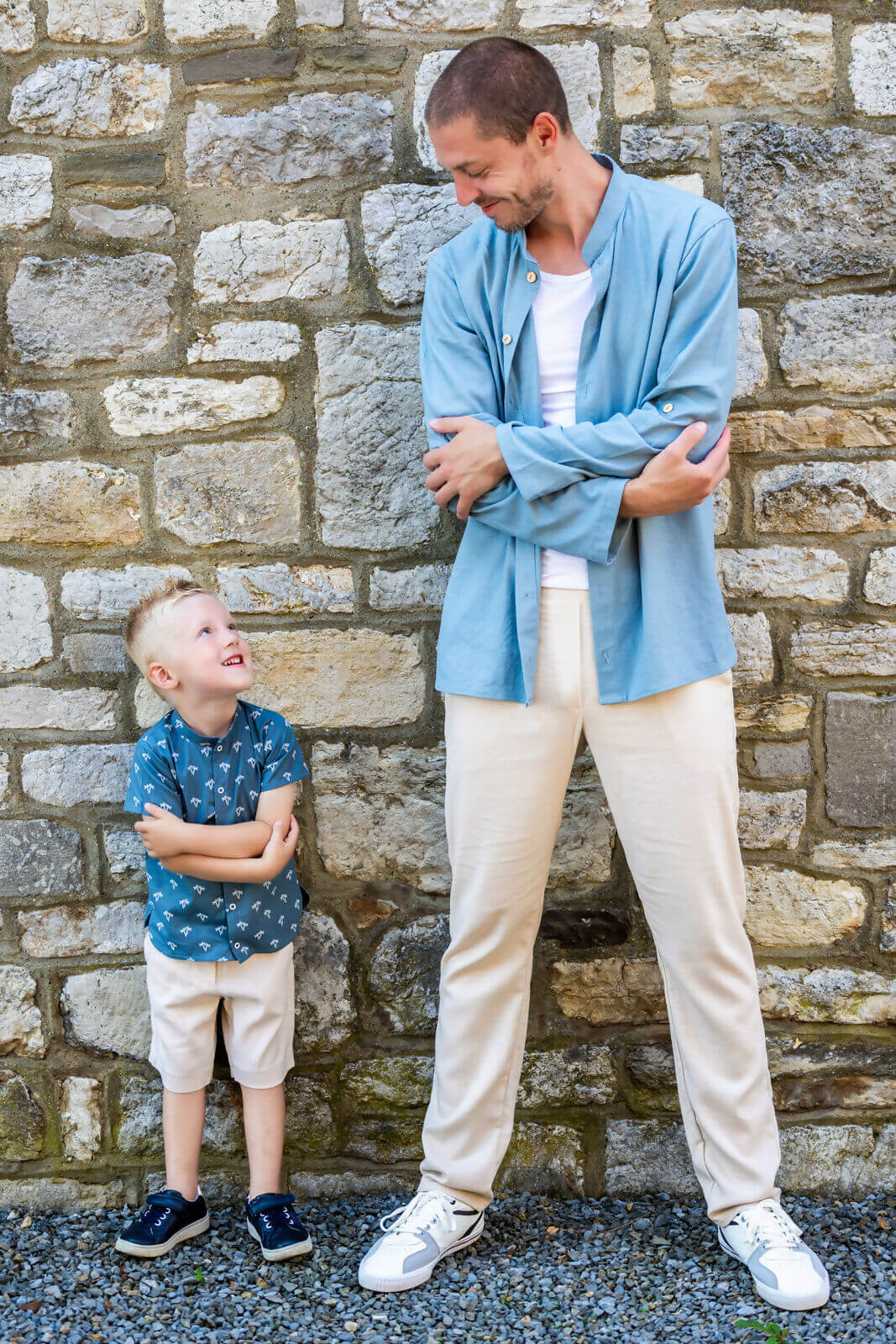 Père en chemise bleue et fils en short beige sourient l'un à l'autre devant un mur de pierre.