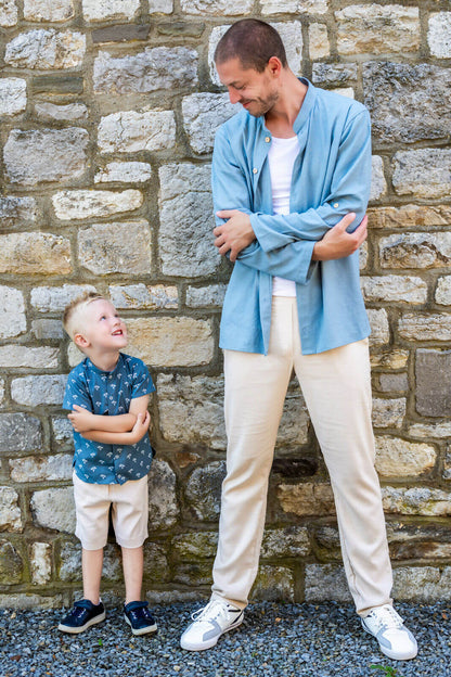 Père en chemise bleue et fils en short beige sourient l'un à l'autre devant un mur de pierre.
