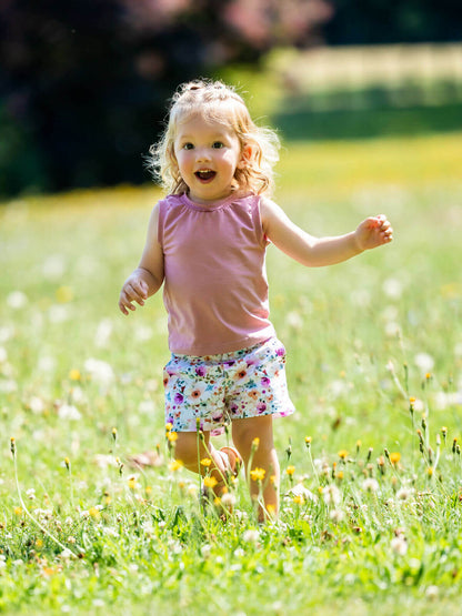Enfant joyeuse portant un short à motif floral, courant dans un champ de fleurs. KUMQUAT - Le short / bermuda / pantalon, patron de couture pdf à télécharger.