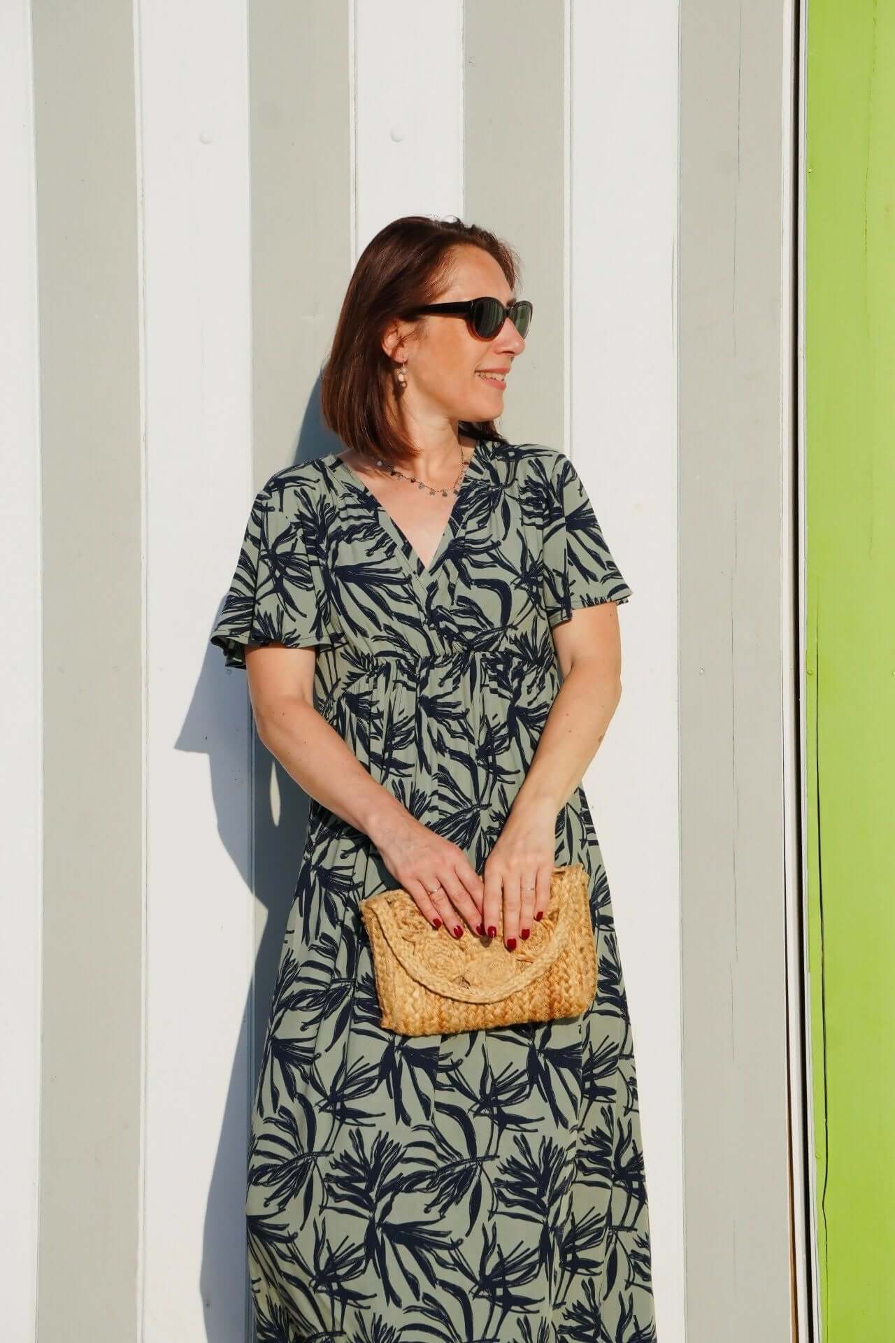 La robe (ou blouse) Sorelle du 32 au 52 portée devant un mur à rayures, style élégant avec sac en osier.