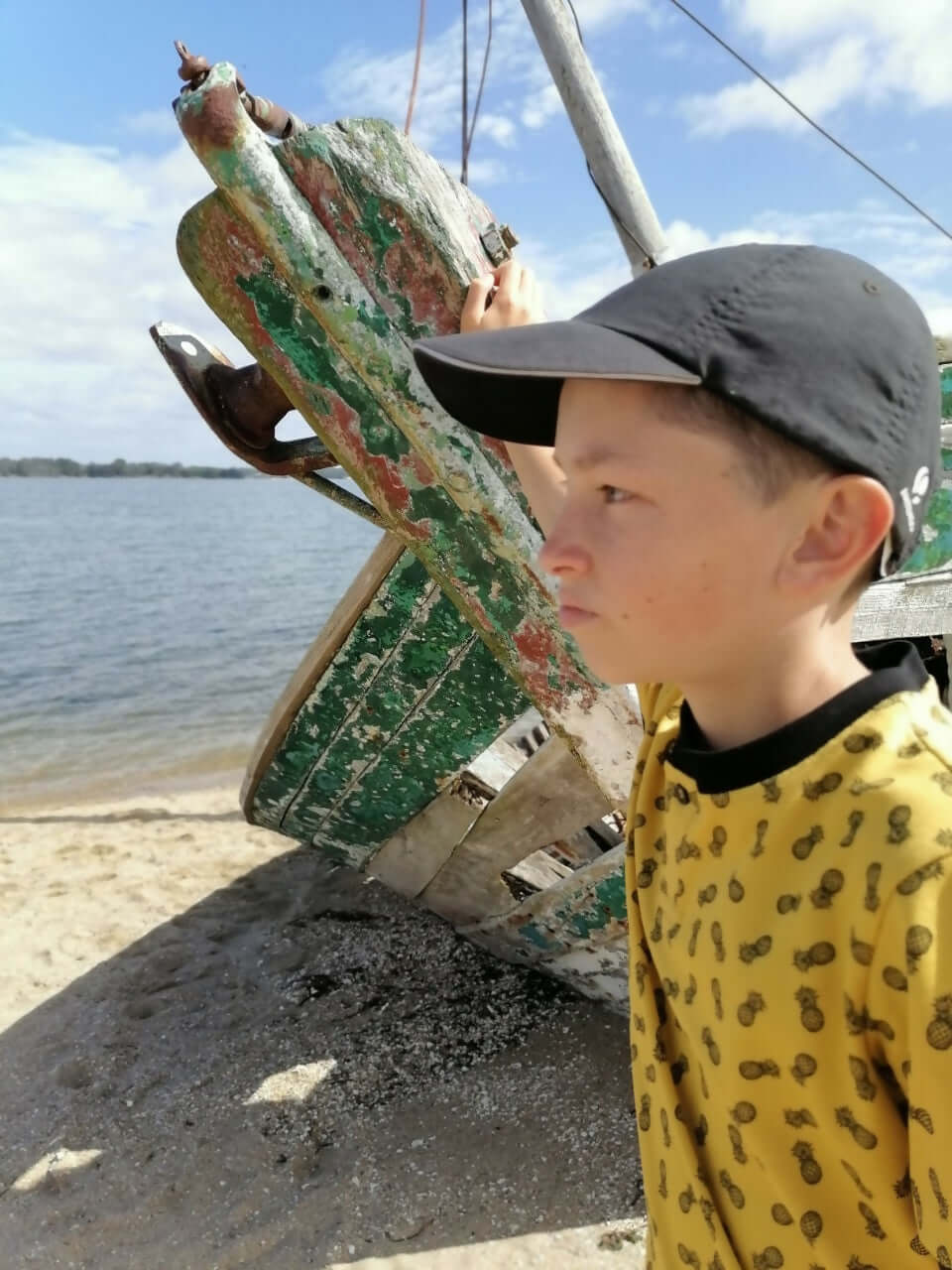 Enfant regardant un bateau en bois usé, vêtu d'un polo jaune à motifs, au bord de l'eau.