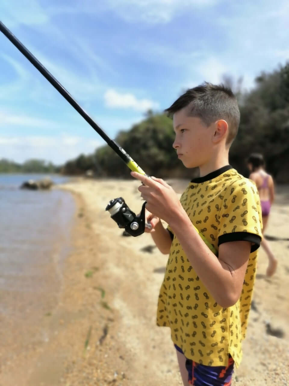 Un garçon pêche sur la plage portant un t-shirt à motifs jaunes, en train de profiter d'une journée ensoleillée.