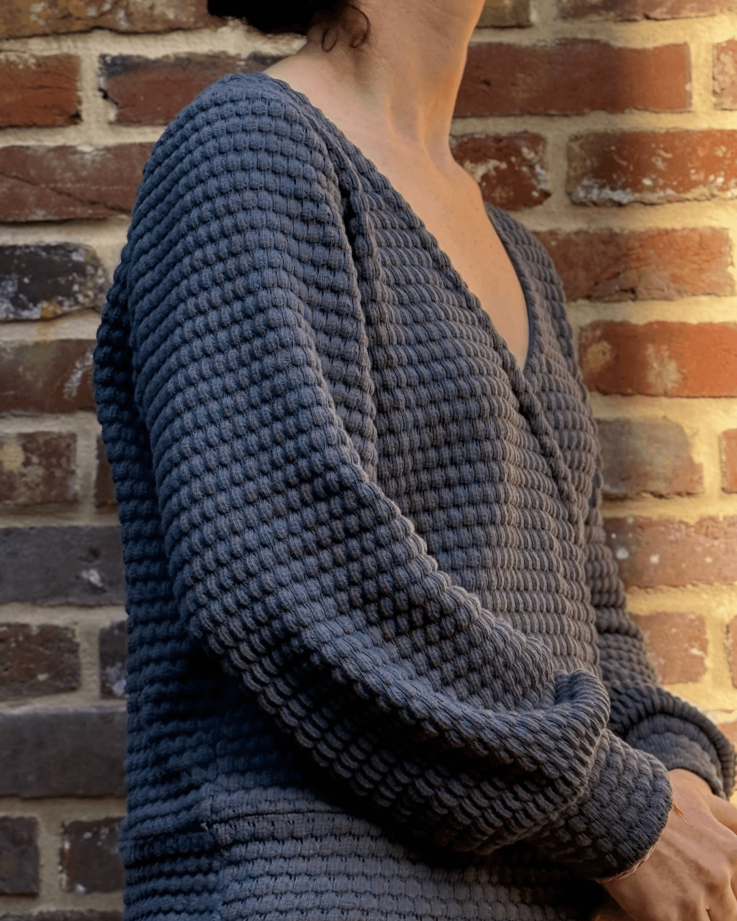 Le pull Axia du 32 au 52 en version cache-cœur, patron de couture pdf à télécharger, contre un mur de briques rustiques.
