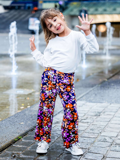 Fille souriante en t-shirt blanc et pantalon fleuri coloré, posant joyeusement près de fontaines.