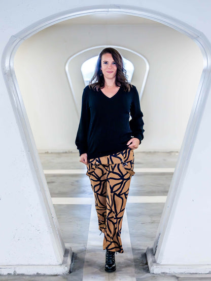 Femme portant un pantalon à motifs feuilles, vêtue d'un pull noir, posant dans un couloir moderne.