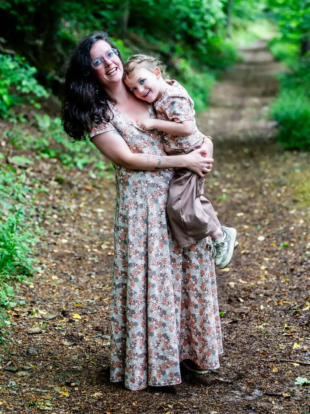 Une mère et son enfant portant des robes assorties florales, souriant dans un chemin boisé.