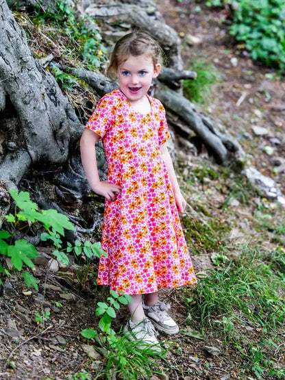 Enfant portant une robe colorée en promenade dans la forêt, souriante près des racines d'un arbre.