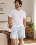 Homme portant le Patron PDF Pyjama NYX, tee-shirt blanc et short rayé bleu et blanc, dans une chambre épurée