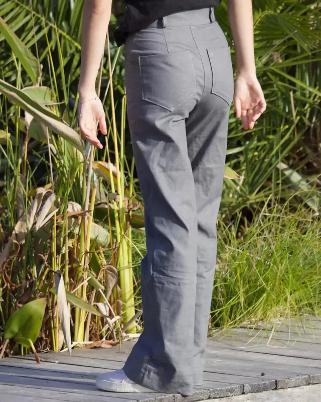 Femme portant le Patron PDF Pantalon Q du 32 au 60, vue de profil en sergé gris, sur une terrasse bois entourée de plantes
