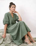 Femme portant le Patron PDF Robe Longue IRIS, manches courtes, en lin, assise sur un lit crème, coupe fluide et élégante