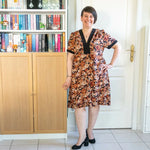 Femme portant le Patron PDF Robe courte Pomme Golden, manches courtes à fleurs orangées et noires, devant une bibliothèque