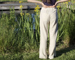 Femme portant le Patron PDF Pantalon A du 32 au 60, taille haute en lin blanc, debout dans un jardin avec iris jaunes