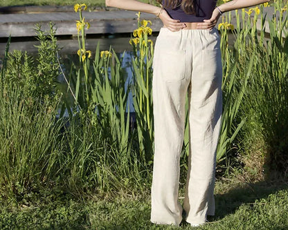 Femme portant le Patron PDF Pantalon A du 32 au 60, taille haute en lin blanc, debout dans un jardin avec iris jaunes