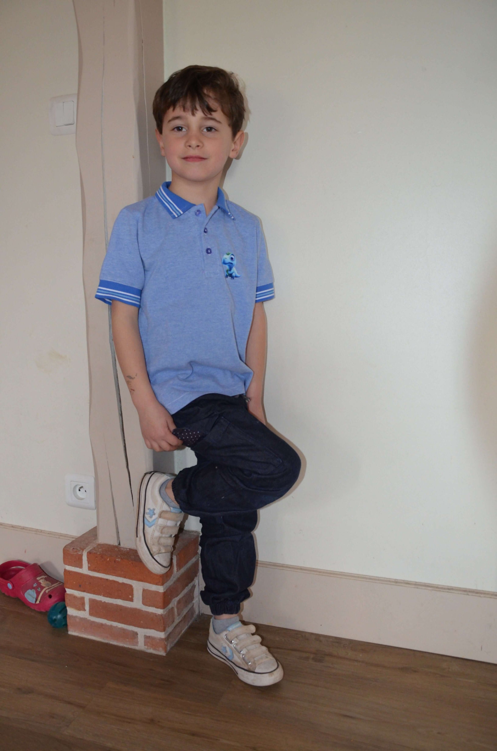 Enfant portant un Polo MARCOLOPO bleu, manches courtes, devant un mur. Patron de couture PDF à télécharger disponible.