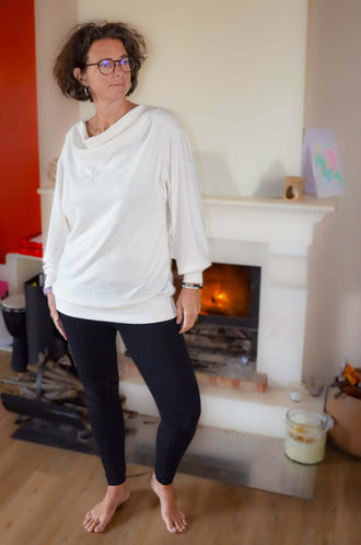 Femme portant un Pull CRYSTAL blanc, manches chauve-souris, col bénitier devant un feu, idéal pour patron de couture pdf à télécharger.