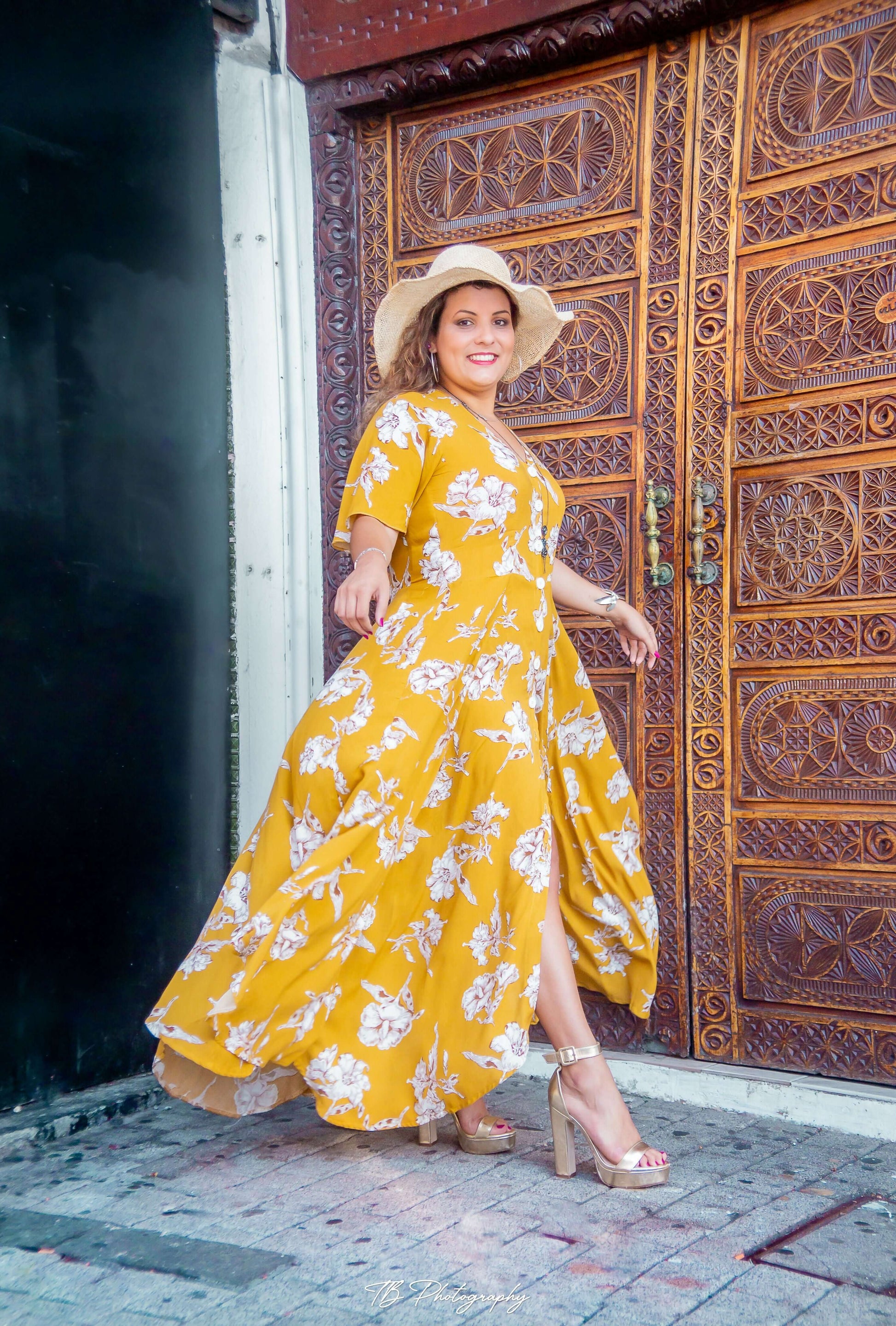 Femme souriante en robe longue jaune fleuri, posant devant une porte en bois ornée.