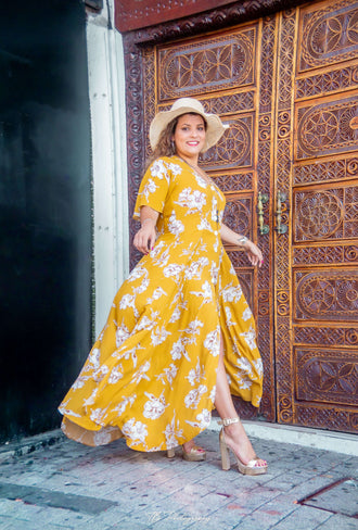 Femme souriante en robe longue jaune fleuri, posant devant une porte en bois ornée.